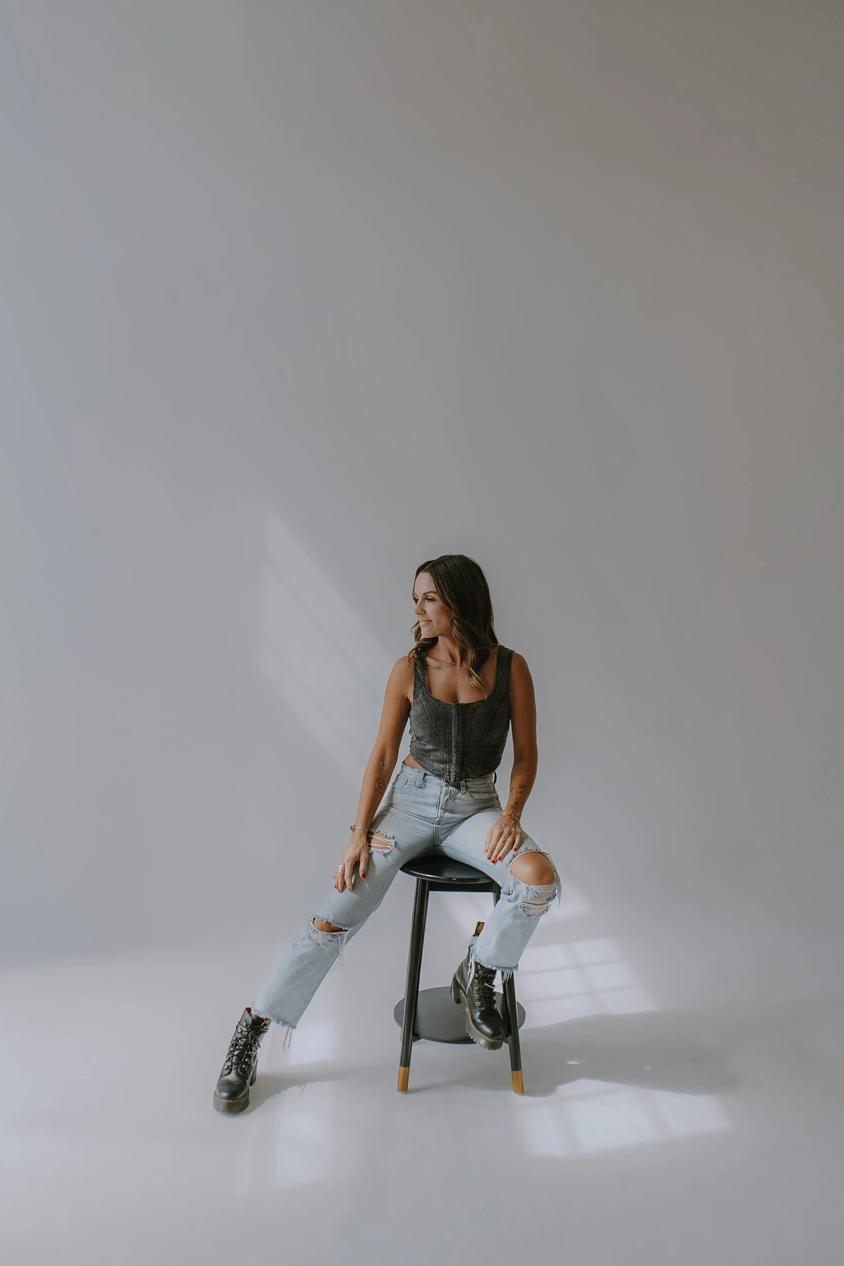 A woman with long brown hair wearing a black sleeveless top, ripped light blue jeans, and black combat boots, sitting on a black stool in a minimalistic studio setting with plain light gray walls.