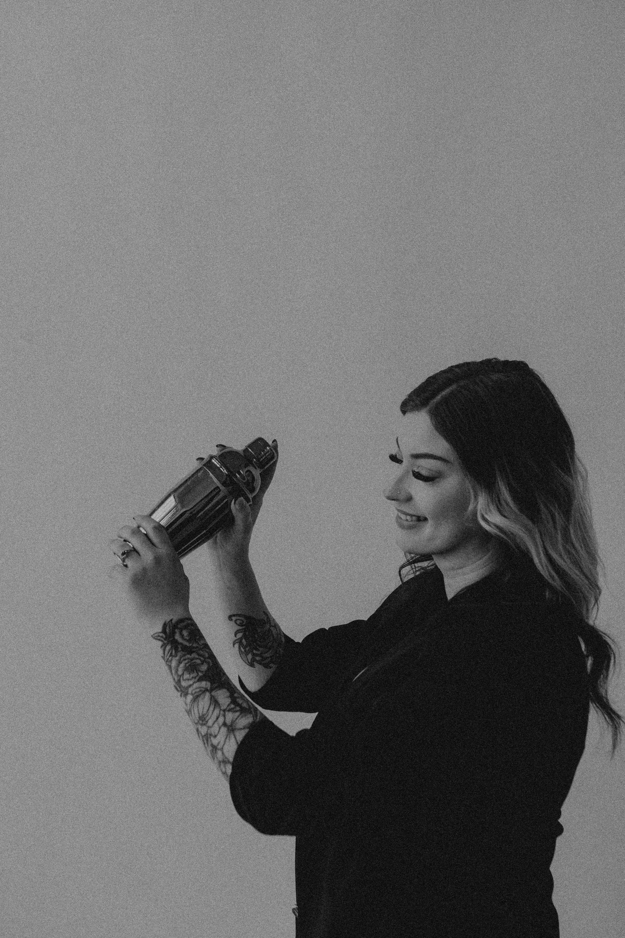 A black and white photo of a woman with long wavy hair smiling while holding a coffee thermos, wearing a dark shirt with tattoos on her arms.