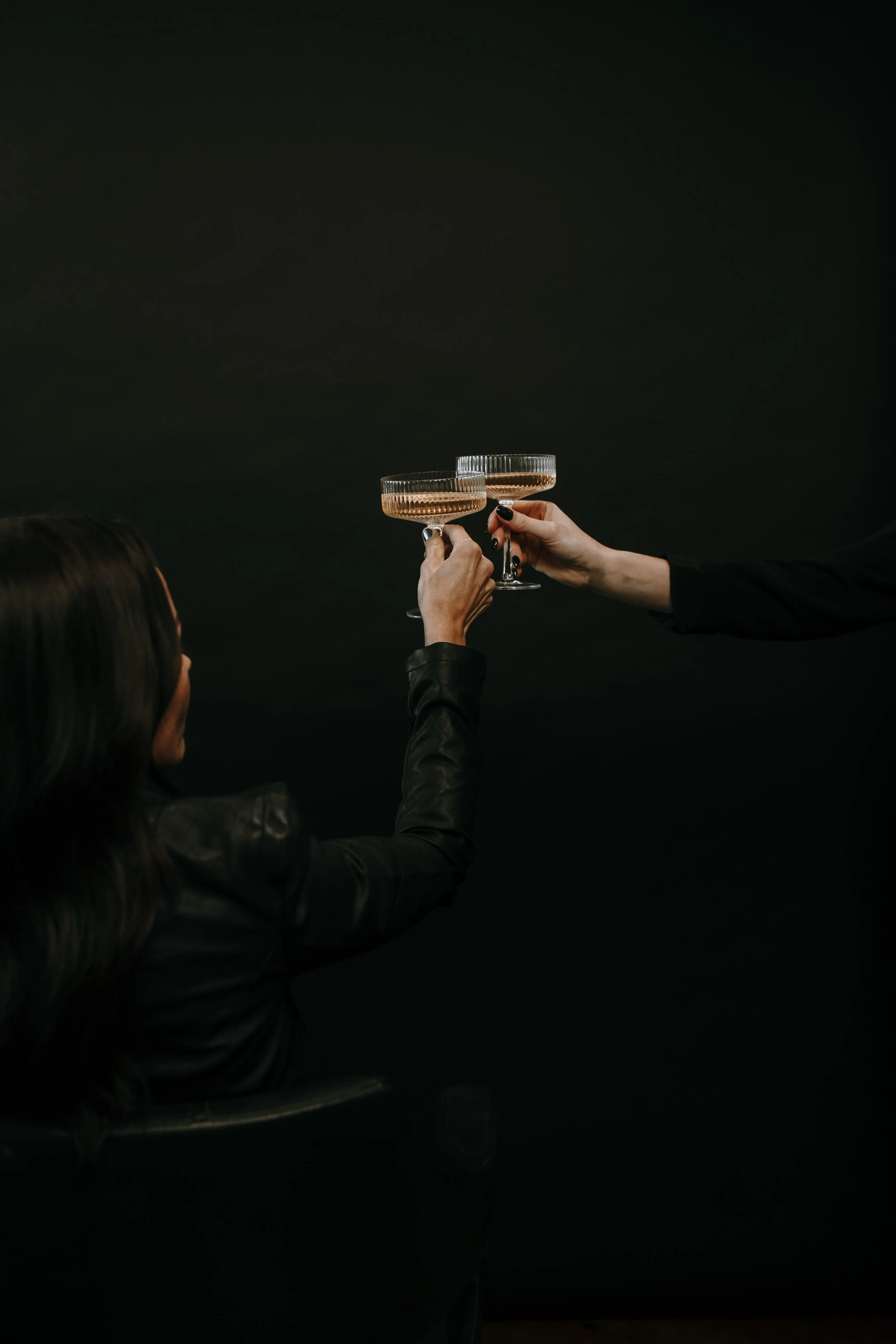Two people wearing black clothing holding glasses of what appears to be champagne or sparkling wine against a dark background.