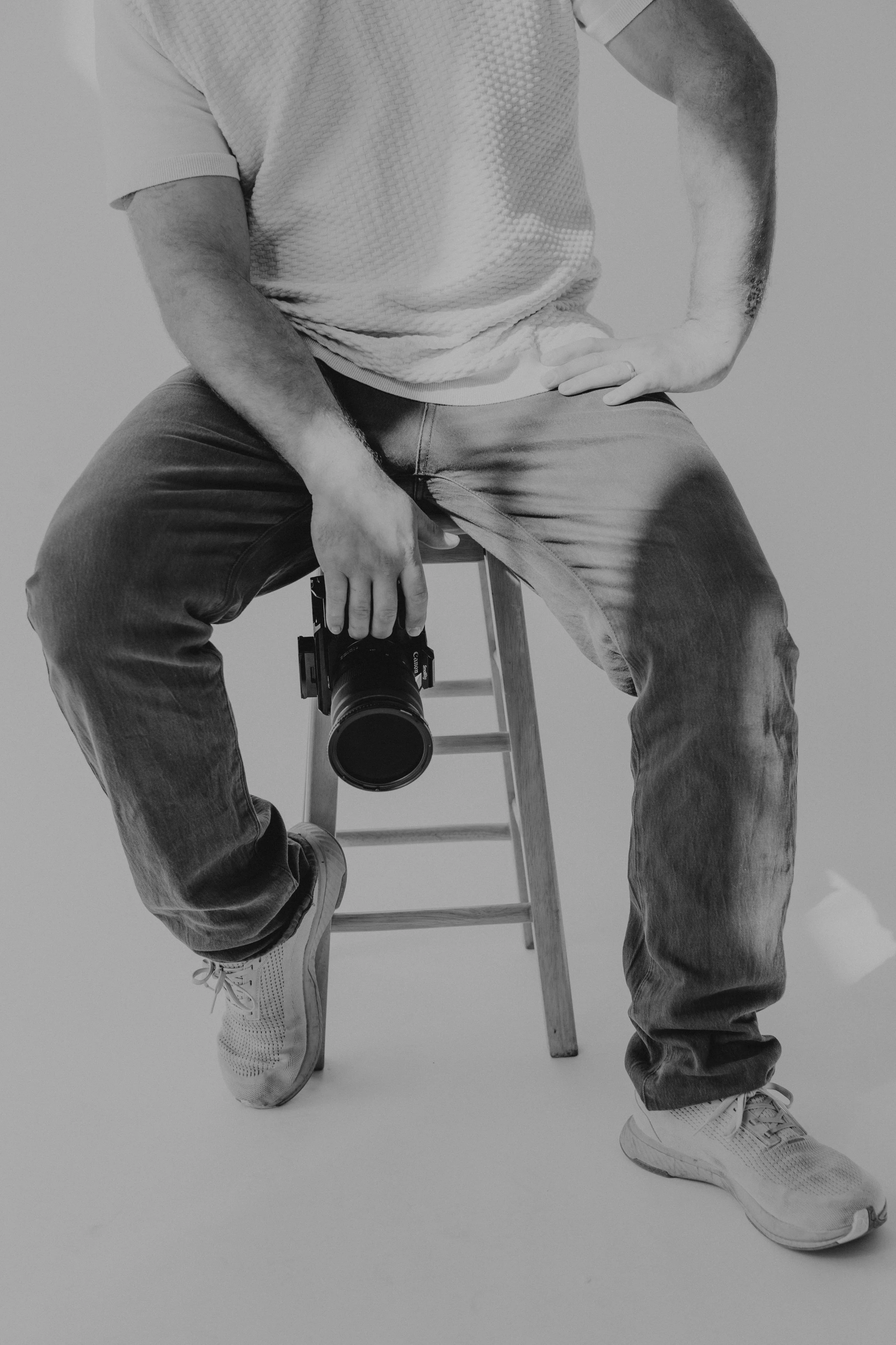 Person sitting on a wooden stool with one hand on their hip and the other holding a camera, wearing a short-sleeved textured shirt, jeans, and sneakers, with one leg crossed over the other, in a black-and-white studio setting.