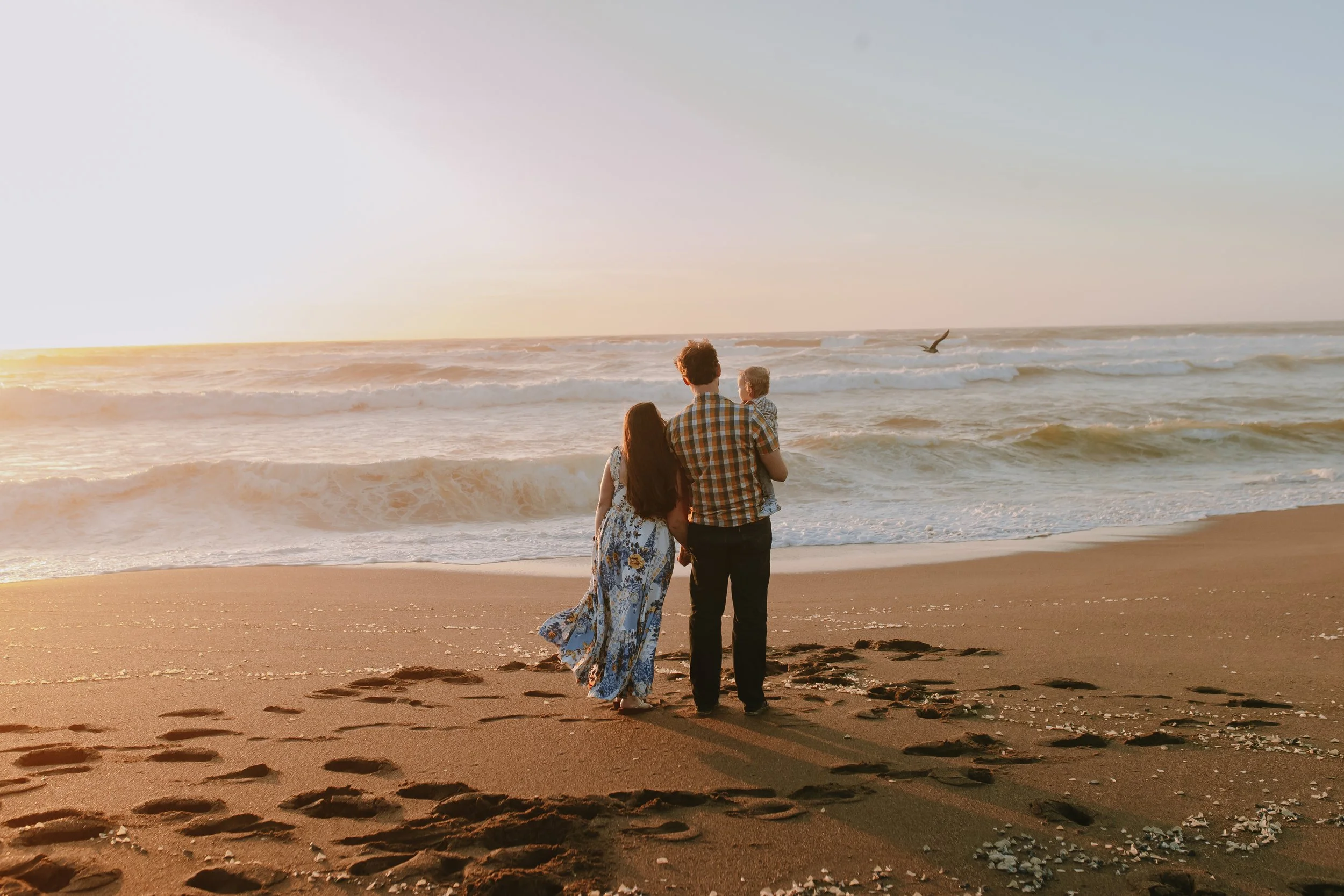 A family of three, a man, a woman, and a small child, standing on a sandy beach and looking out at the ocean during sunset.