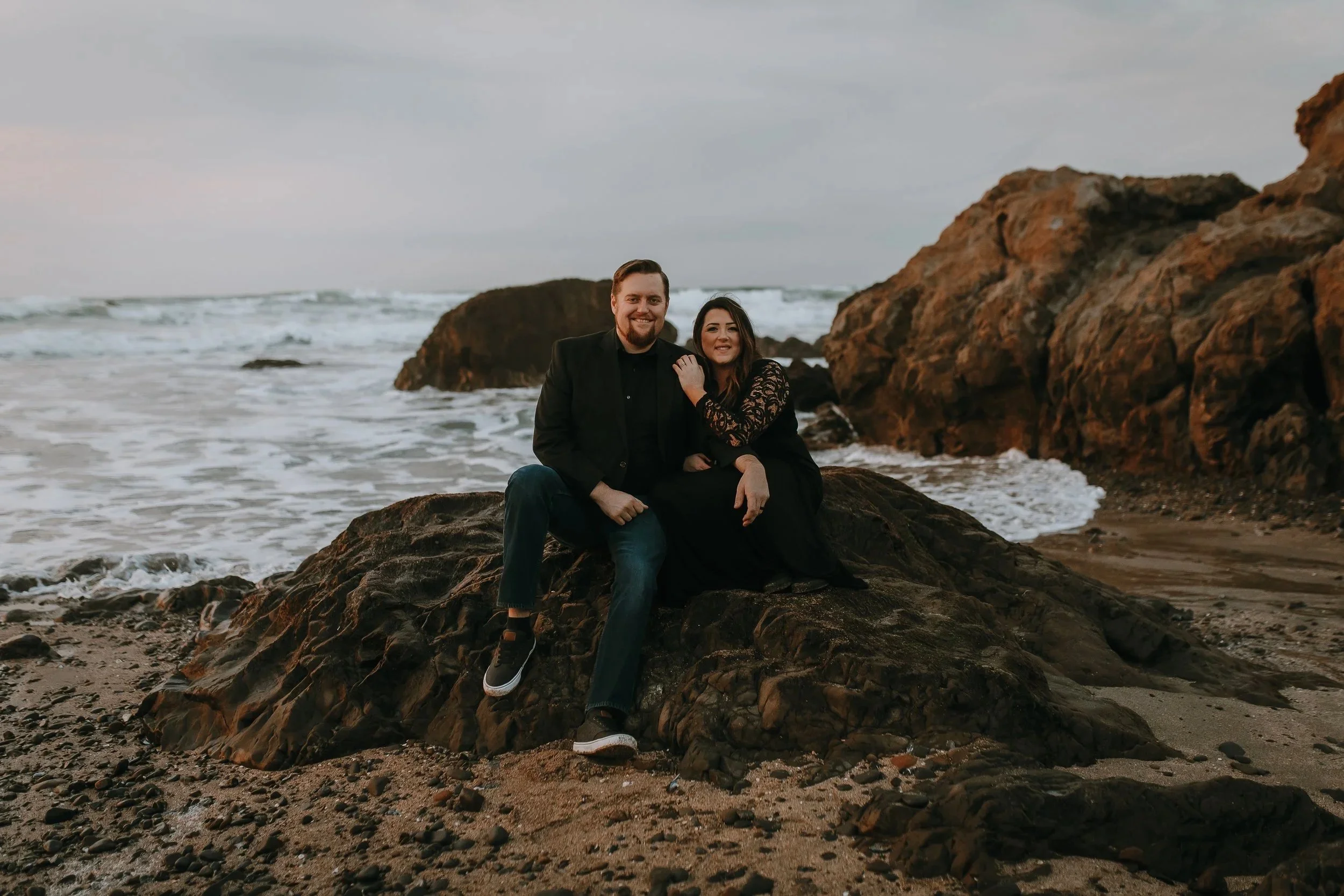 A smiling couple sitting on rocks at the beach near large rock formations, with waves and the ocean in the background during a cloudy day.