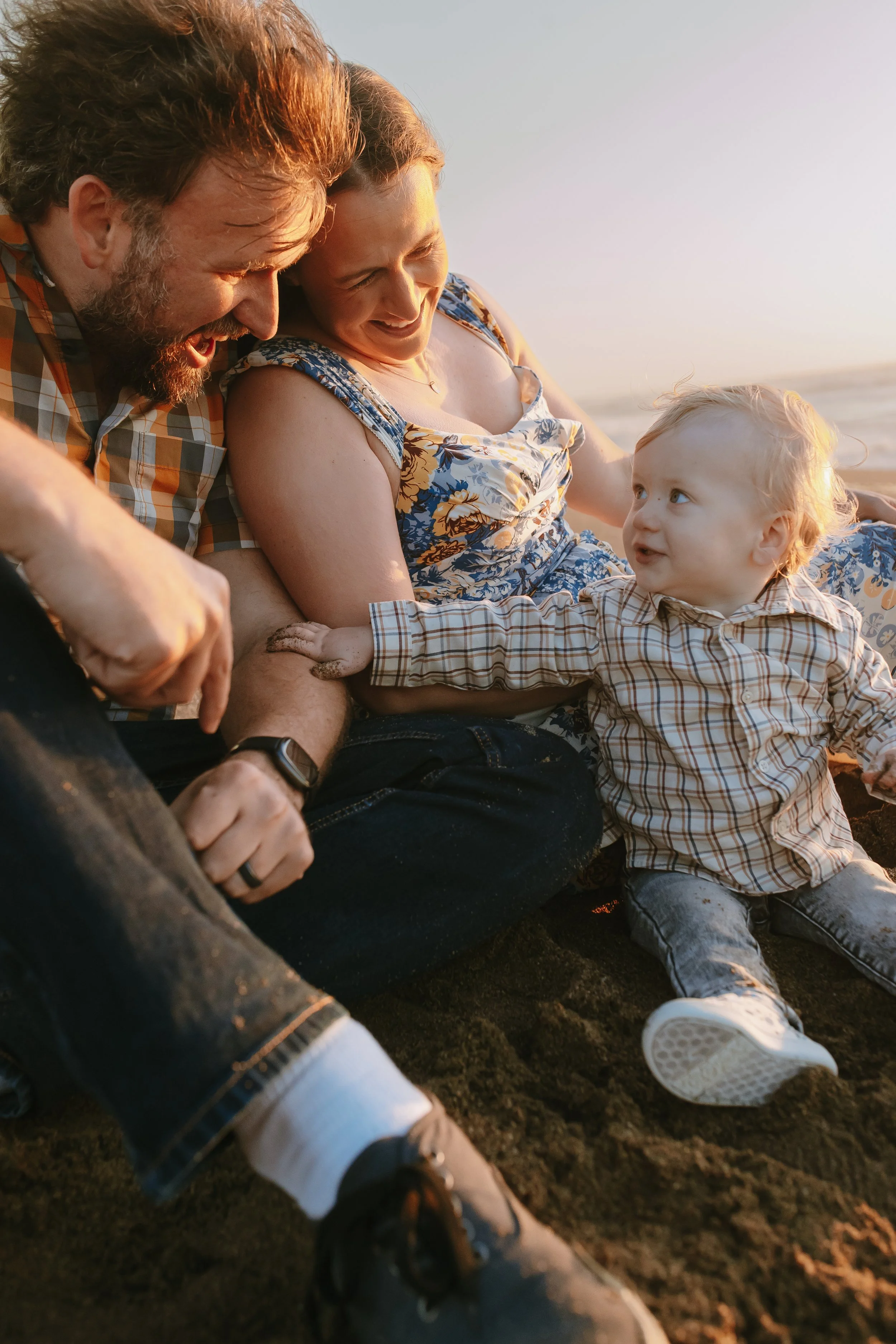 A family of three sitting together on a sandy beach at sunset, smiling and looking at each other.