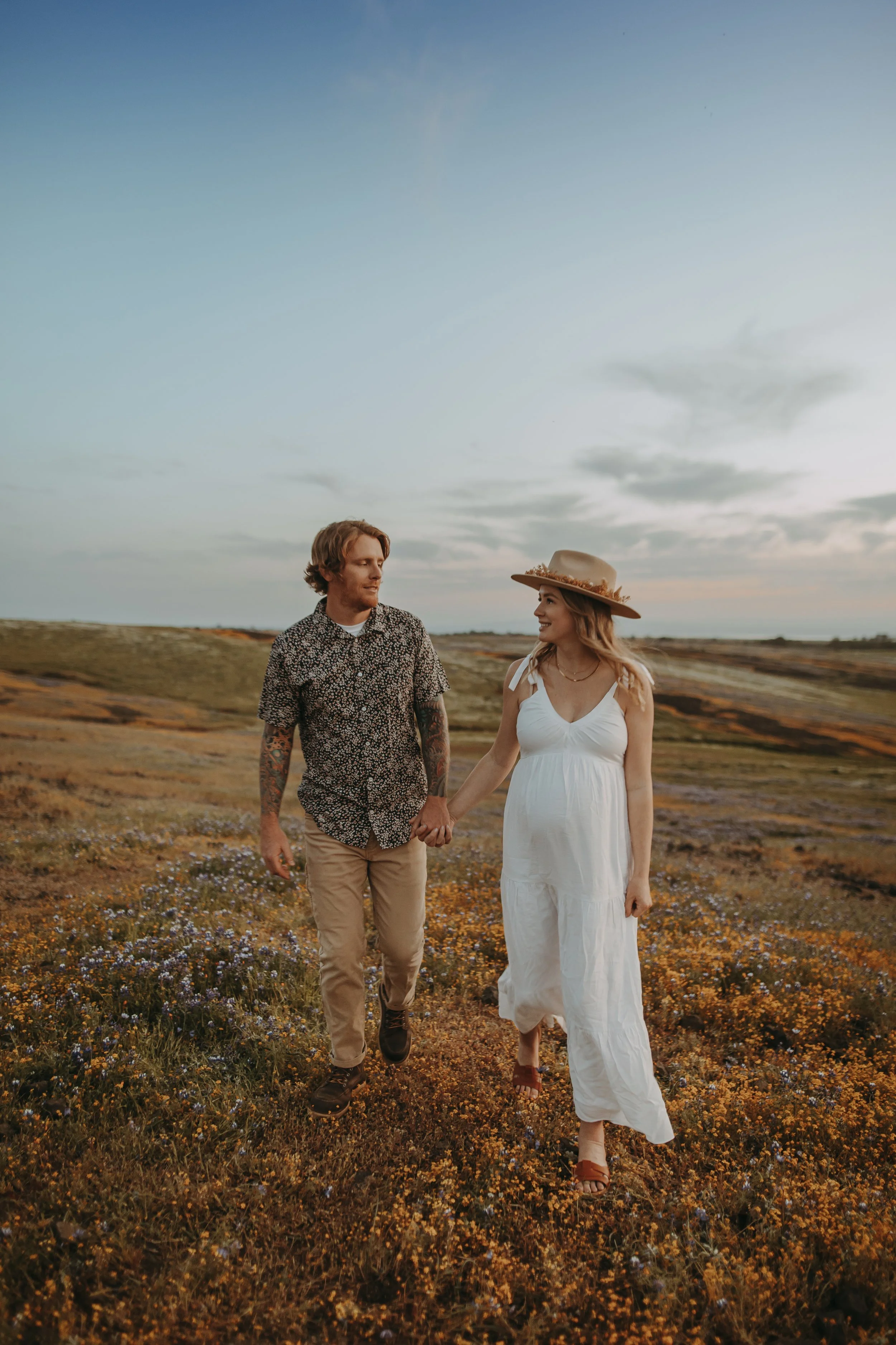 A couple holding hands and walking through a field of yellow and purple wildflowers during sunset. The woman is wearing a white dress and a wide-brimmed hat, while the man is dressed in a patterned shirt and khaki pants. The sky is partly cloudy with a calm, serene atmosphere.