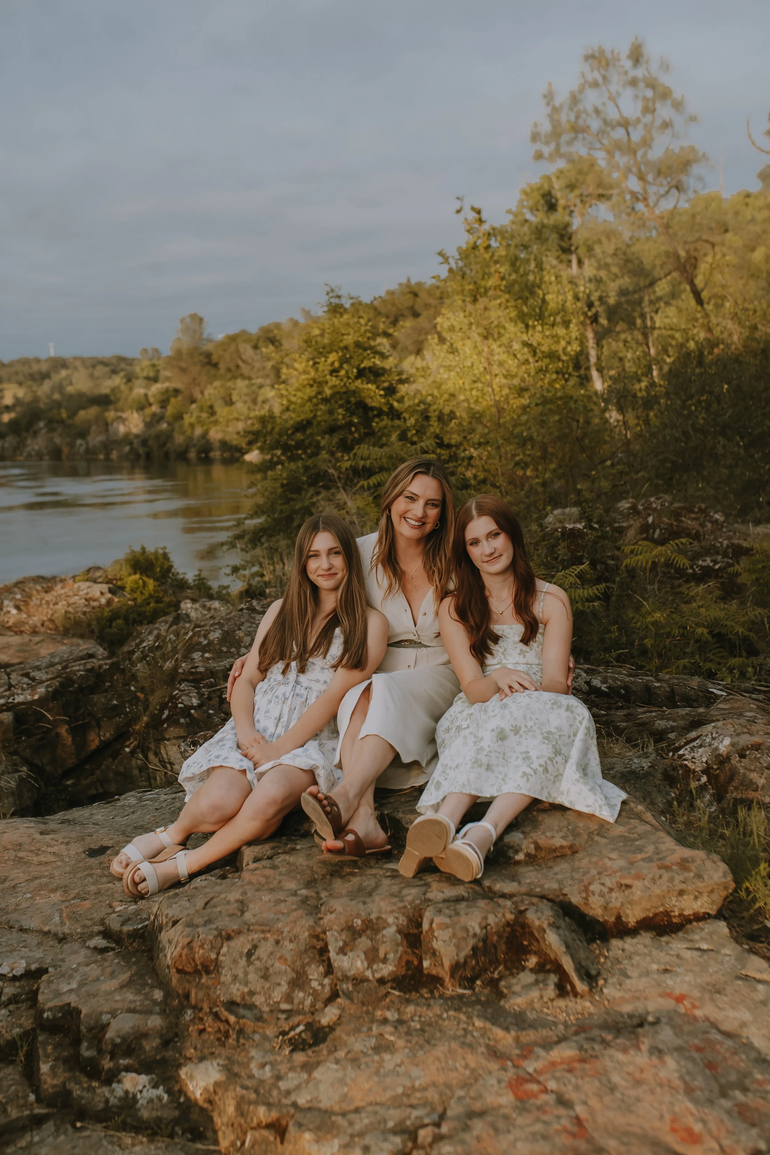 A woman and two young girls sitting on rocks by a river in a forested area, smiling at the camera, during sunset or late afternoon.
