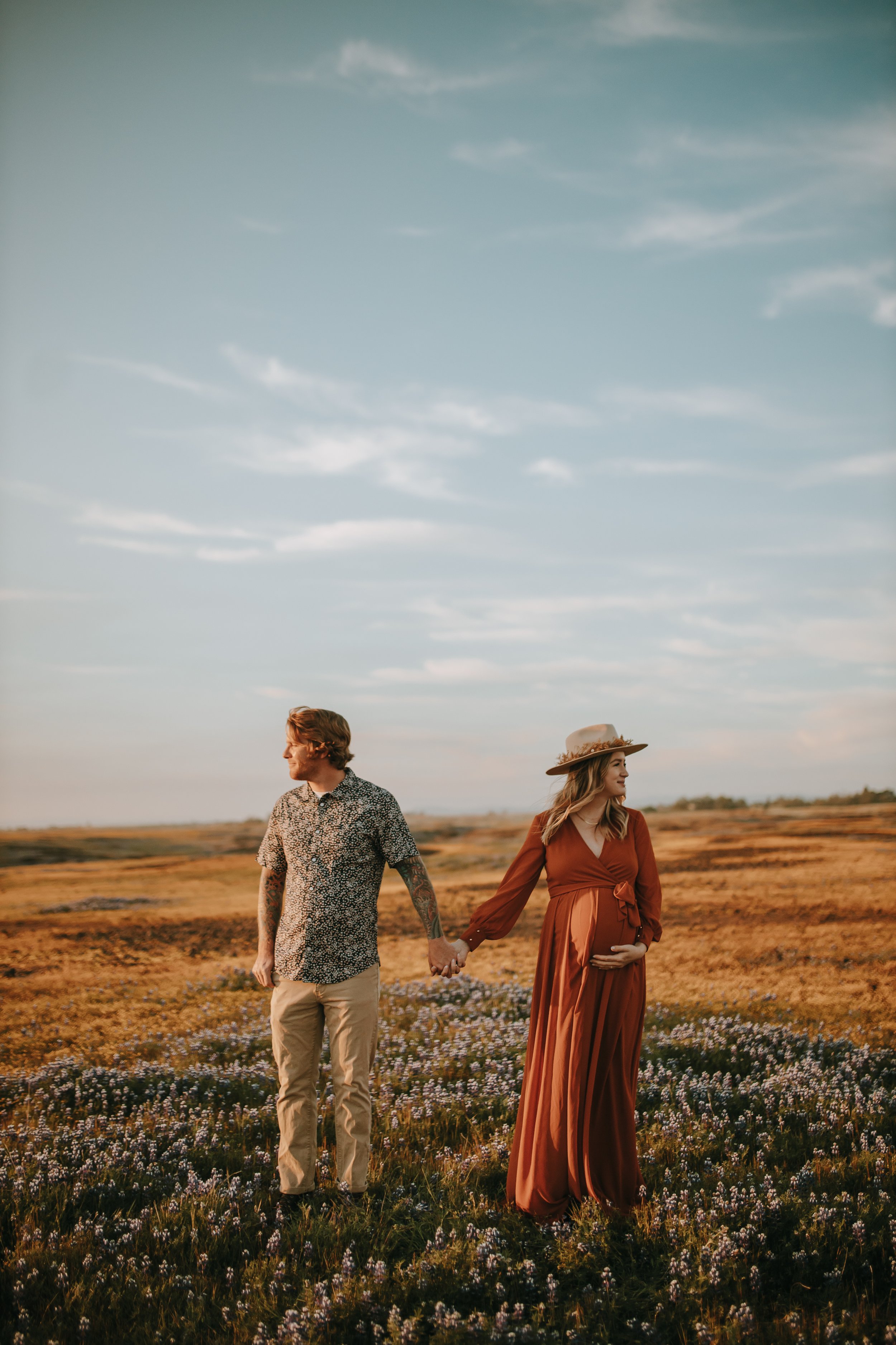 A pregnant woman in a long rust-colored dress holding hands with a man in a patterned shirt in a field of purple flowers at sunset.
