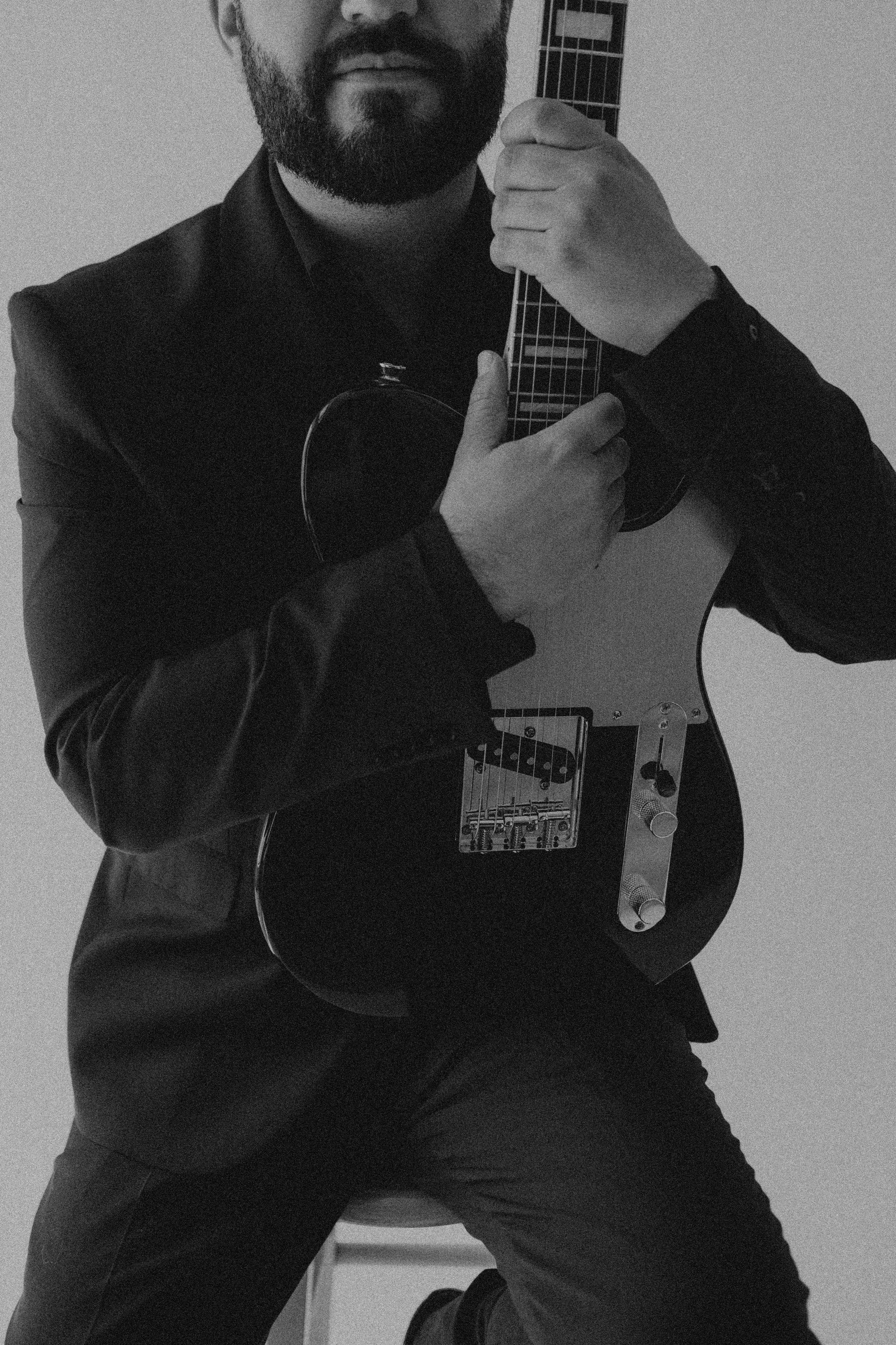 Black and white photo of a man with a beard holding an electric guitar, wearing a jacket and dark pants, sitting on the floor in front of a plain wall.