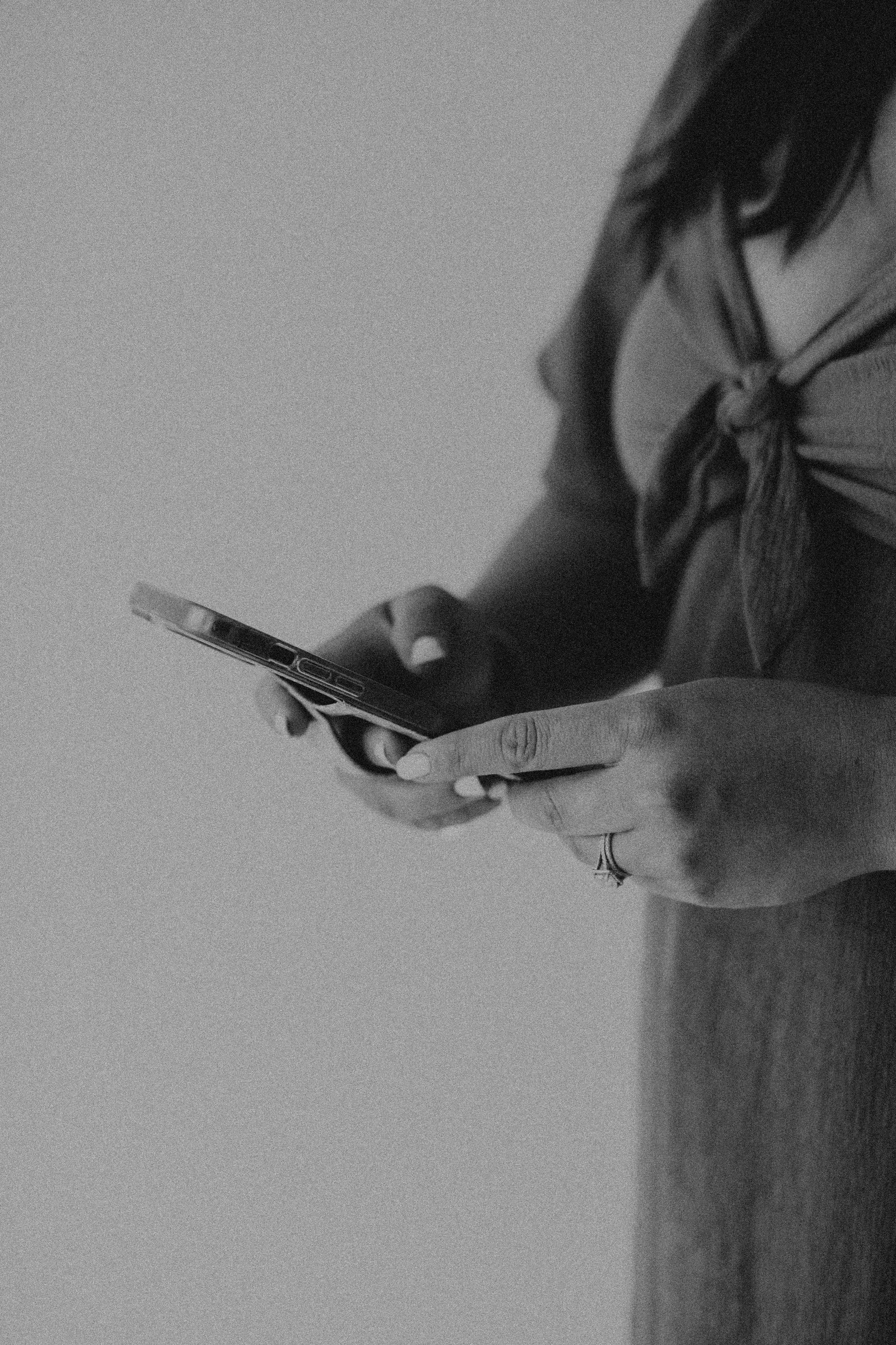 A person holding a smartphone in their right hand, wearing a ring on their right middle finger, with a dark-colored jacket and a striped scarf. The image is in black and white.