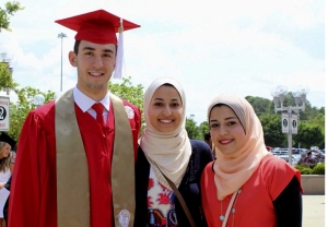 Photo Description: Two women stand center and right with a man in his red graduation cap and gown. All three are wearing large smiles and you can see happiness in their eyes. It seems they are in a parking lot, post-graduation ceremony, with some trees, lights, signs, and cars in the background on a bright but cloudy day.