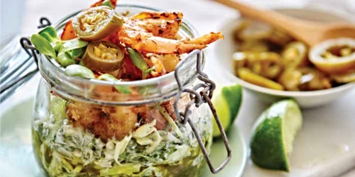 Photo Description: A glass jar is center left, with a metal lid clam, top swung open. In the jar from bottom up, salsa verde, cabbage, grilled shrimp, sunflower sprouts, and jalapeno. in a blur on a plate to the left a couple lime wedges, and behind that even more blurred, a small bowl of jalapenos with a wooden spoon.