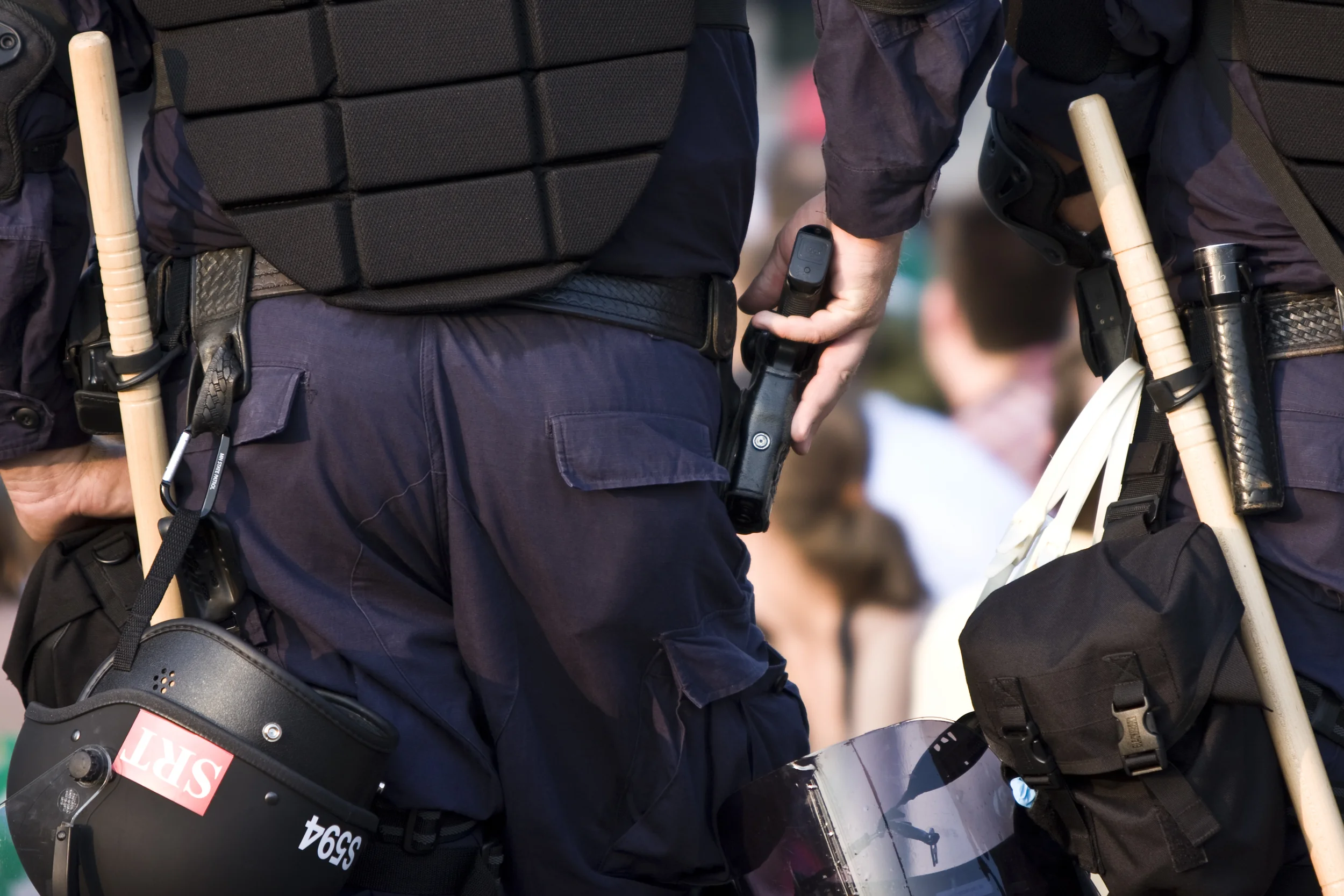 Photo Description: A rear-view of two police officers standing before a blurry crowd. The officer to the left has his hand on his gun. They are both equiped with weapons, armor, and helmets.