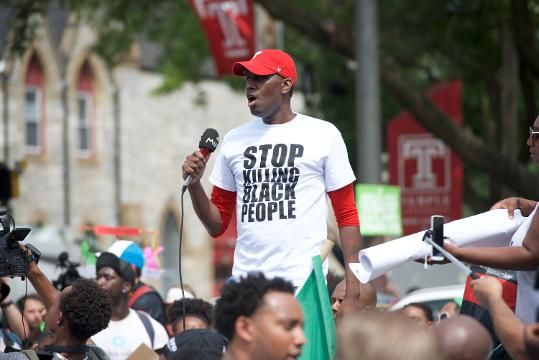 Photo Description: A man stands center of photo wearing a white t-shirt that states "STOP KILLING BLACK PEOPLE." He holds and speaks into a microphone and appears to be standing on a soap box, or something lifting him about 3 feet above the blurry crowd that surrounds him. Photo: Jasper Colt/USA TODAY NETWORK)