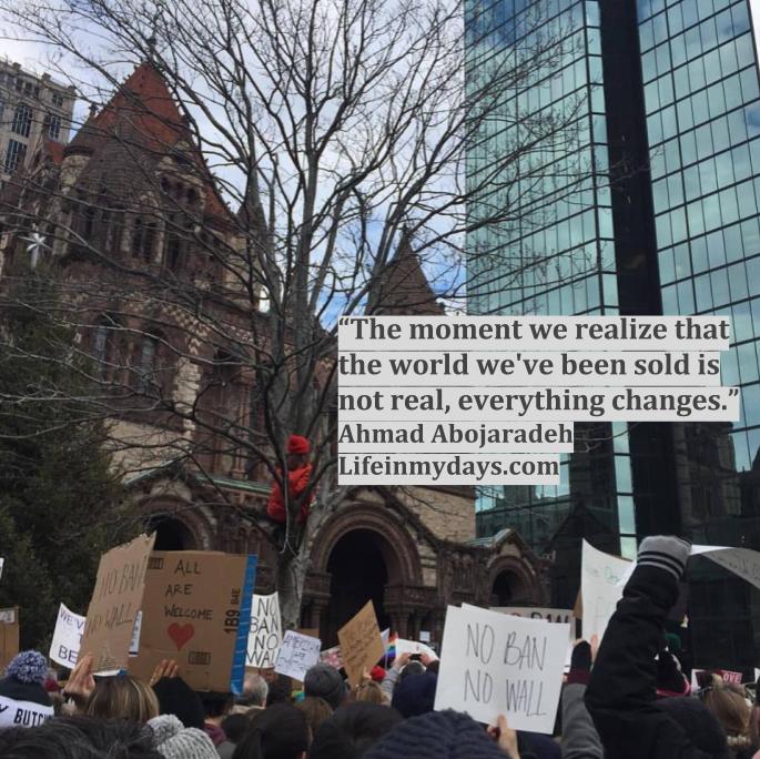 Photo Description: A group of protestors gather in Copley Square in Boston. They carry signed 'NO BAN NO WALL" "ALL ARE WELCOME" and others. Hovering above them is the quote "The moment we realize that the world we've been sold is not real, everything changes."