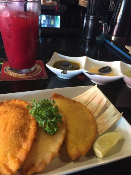 Photo Description: Three crisp fried samosas topped with a large piece of parsley, atop a dried corn husk, with a lime garnish in the forefront. Behind, to the right three dishes containing little spoons and various sauces. To the left behind, a dark red beverage with ice and a straw in a pint glass.