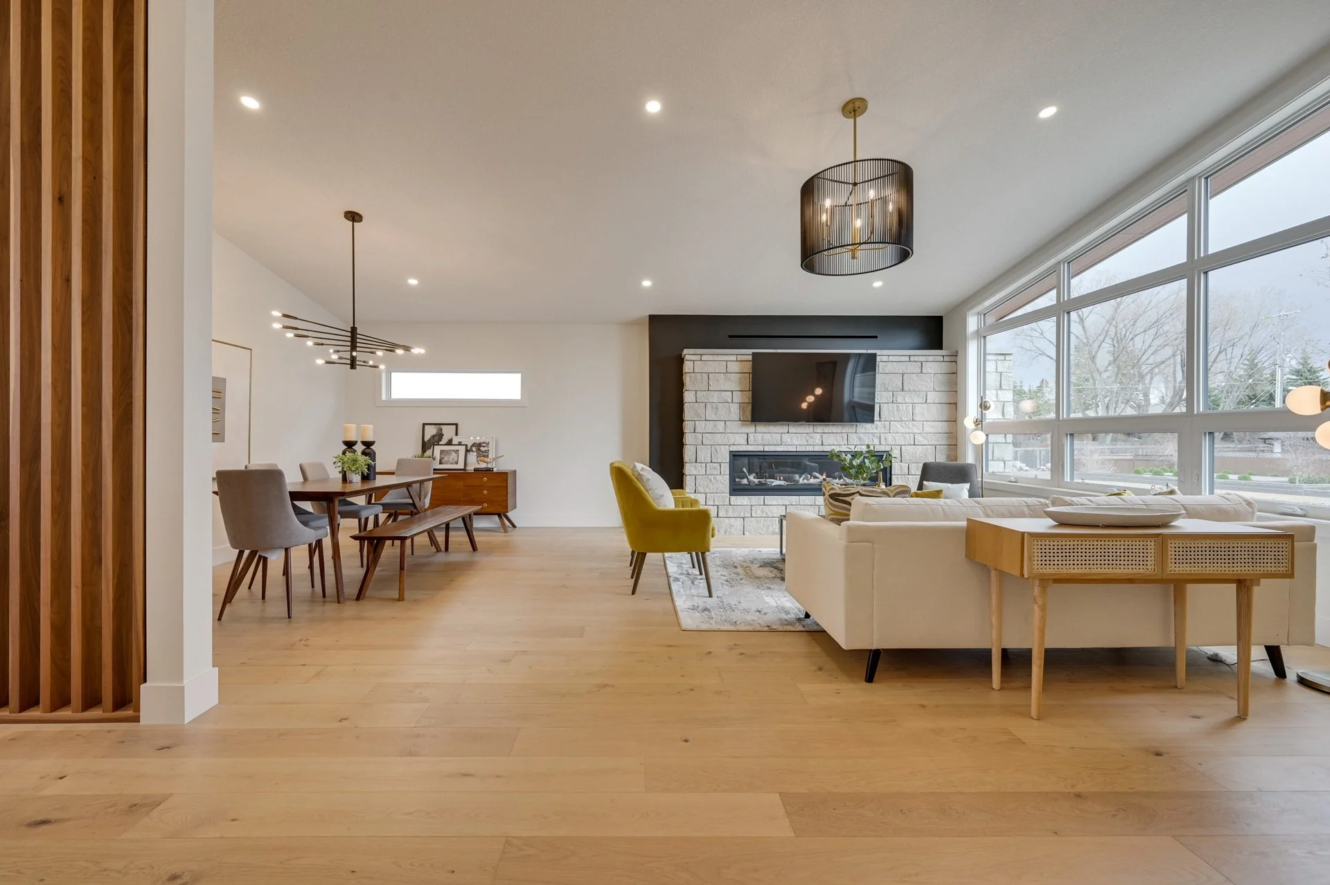 Open-concept living room and dining area with wooden floors, a cream sofa, yellow accent chair, black fireplace, and large windows.
