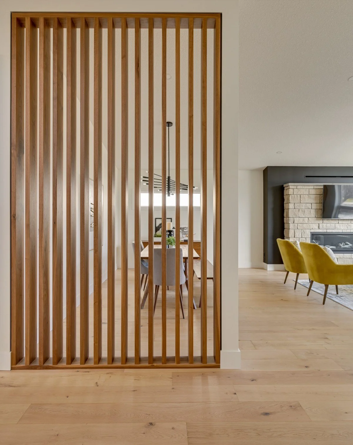 Living and dining rooms separated by a wooden slat partition, hardwood floors, a fireplace, yellow chairs, and a dining table with chairs.