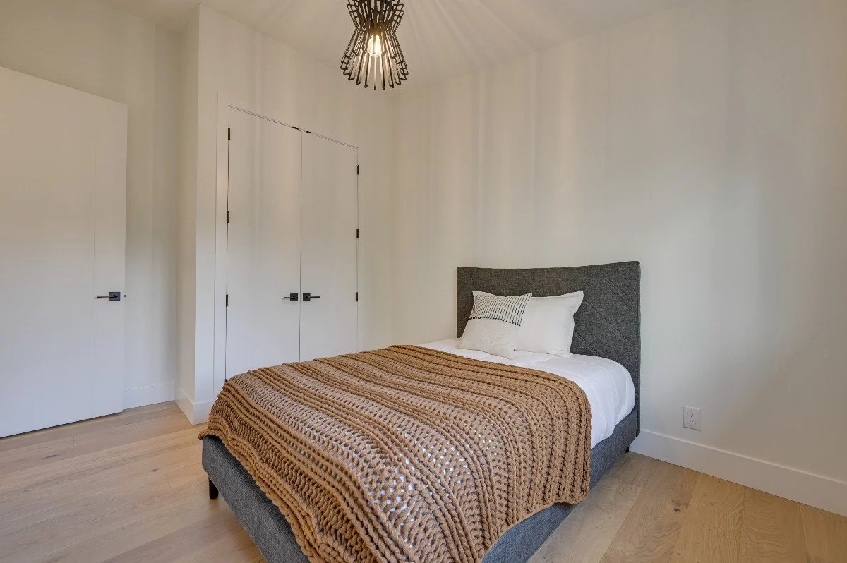 A minimalist bedroom with a bed featuring a gray upholstered headboard, white bedding, and a brown textured throw blanket. There are two pillows, one striped and one plain. The room has light wooden floors, a white wall, a ceiling light fixture with 