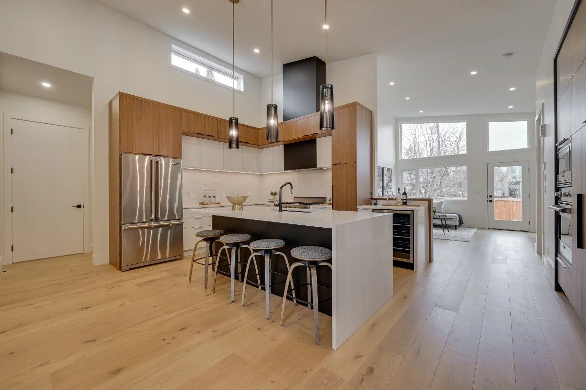 Modern kitchen with light wood flooring, white walls, and large windows, featuring a central island with bar stools, stainless steel refrigerator, and black cabinetry.