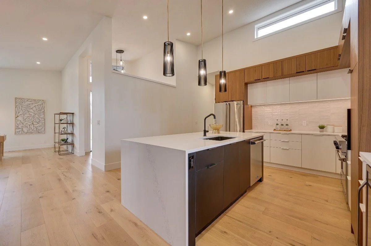 Modern open-concept kitchen with white and wood cabinets, a large white island with a sink, black pendant lights, stainless steel appliances, and light wood flooring.
