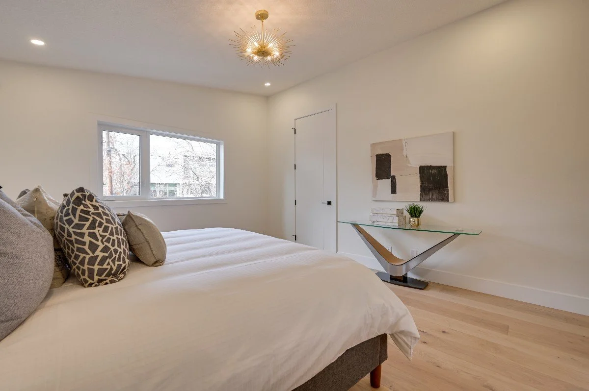 A bedroom with white walls, a large window, and a bed with beige and patterned pillows. There is a modern glass and wood console table against the wall with a plant and decorative objects. A gold starburst ceiling light fixture is above, and the floo
