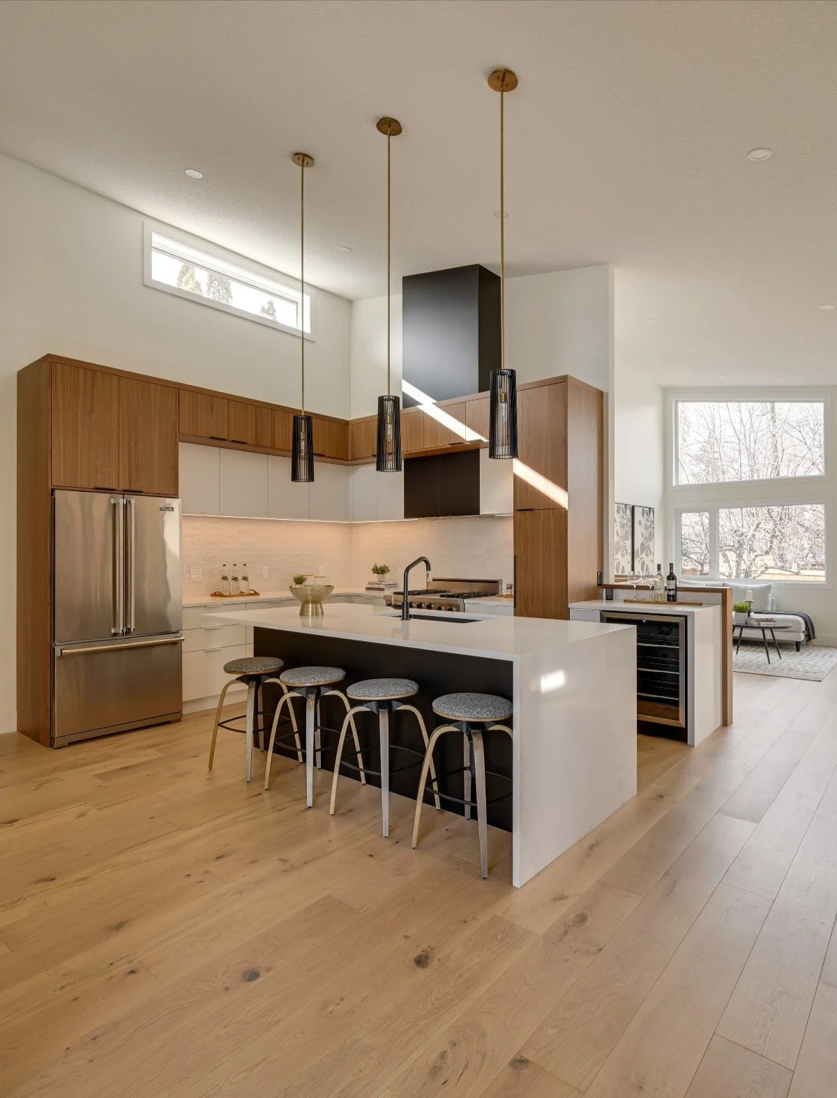 Modern kitchen with a white island, four barstools, wooden cabinets, stainless steel refrigerator, black chimney, and large windows with a living room visible in the background.