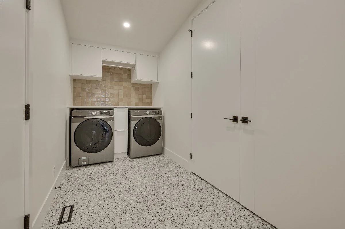A laundry room with a front-loading washer and dryer side by side, white cabinets above, beige backsplash tiles, white walls, and tile flooring.