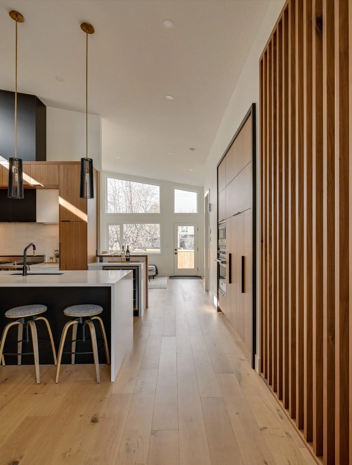 Modern kitchen with wooden accents, black and white cabinetry, a white island with bar stools, pendant lighting, and large windows at the back letting in natural light.