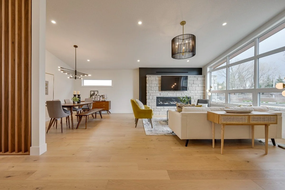 Open-concept living and dining room with wood flooring, a fireplace, large windows, and modern furniture including a white sofa, yellow armchair, dining table and chairs, and decorative elements.