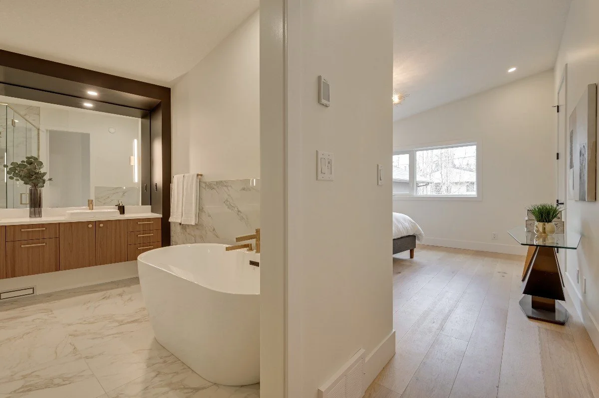 Split view of a modern bathroom on the left with a bathtub, marble floor, wooden vanity, and large mirror; on the right, a bedroom with a large window, light-colored wooden flooring, and minimal decor.
