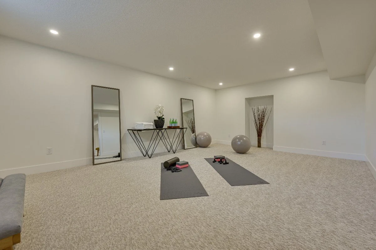 Home gym with two yoga mats, yoga blocks, foam rollers, and weights on a beige carpeted floor, with large mirrors leaning against the wall and exercise balls, decorative vases, and a table with towels on it in the background.