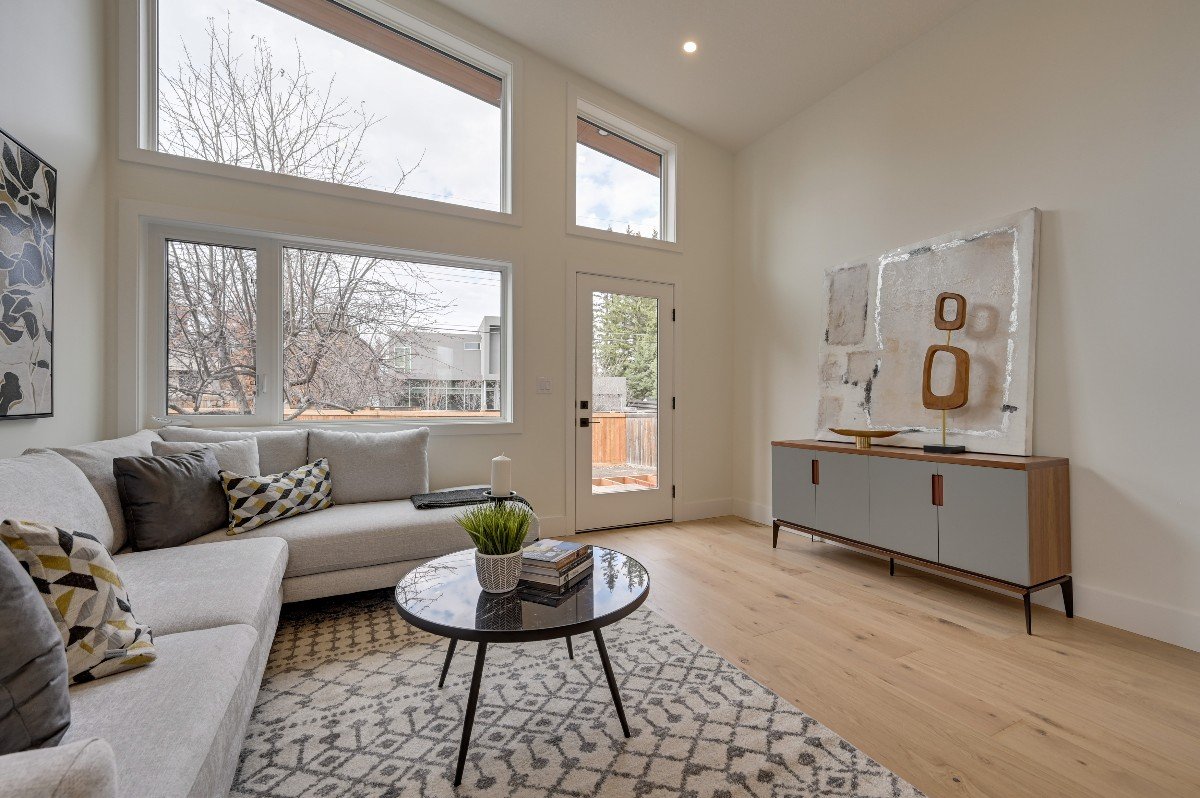 Modern living room with large windows, white walls, a beige sofa with multiple cushions, a round black coffee table with books and a plant, a patterned rug, a sideboard with artistic decor, and a door leading outside.