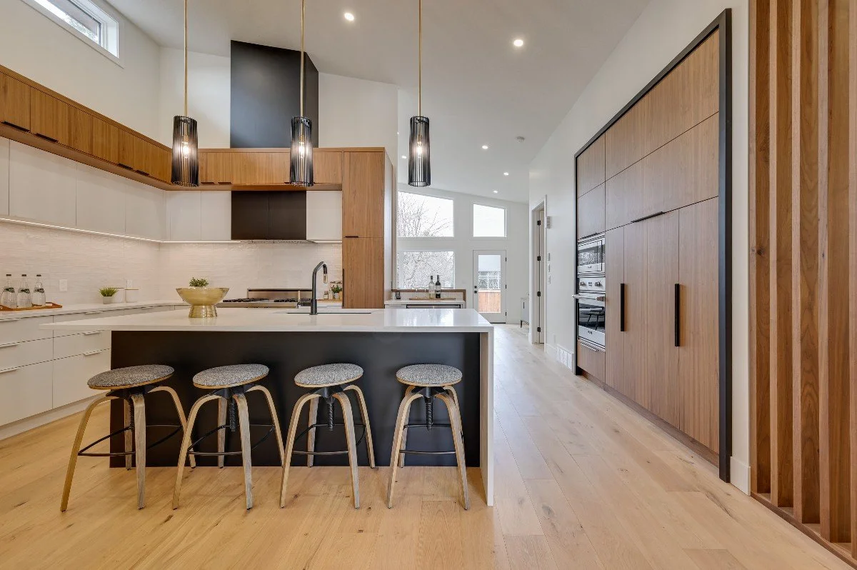 Modern kitchen with a black and white color scheme, featuring four gray stools at a black island with a white countertop, wooden upper and lower cabinets, black pendant lights, and large windows allowing natural light.