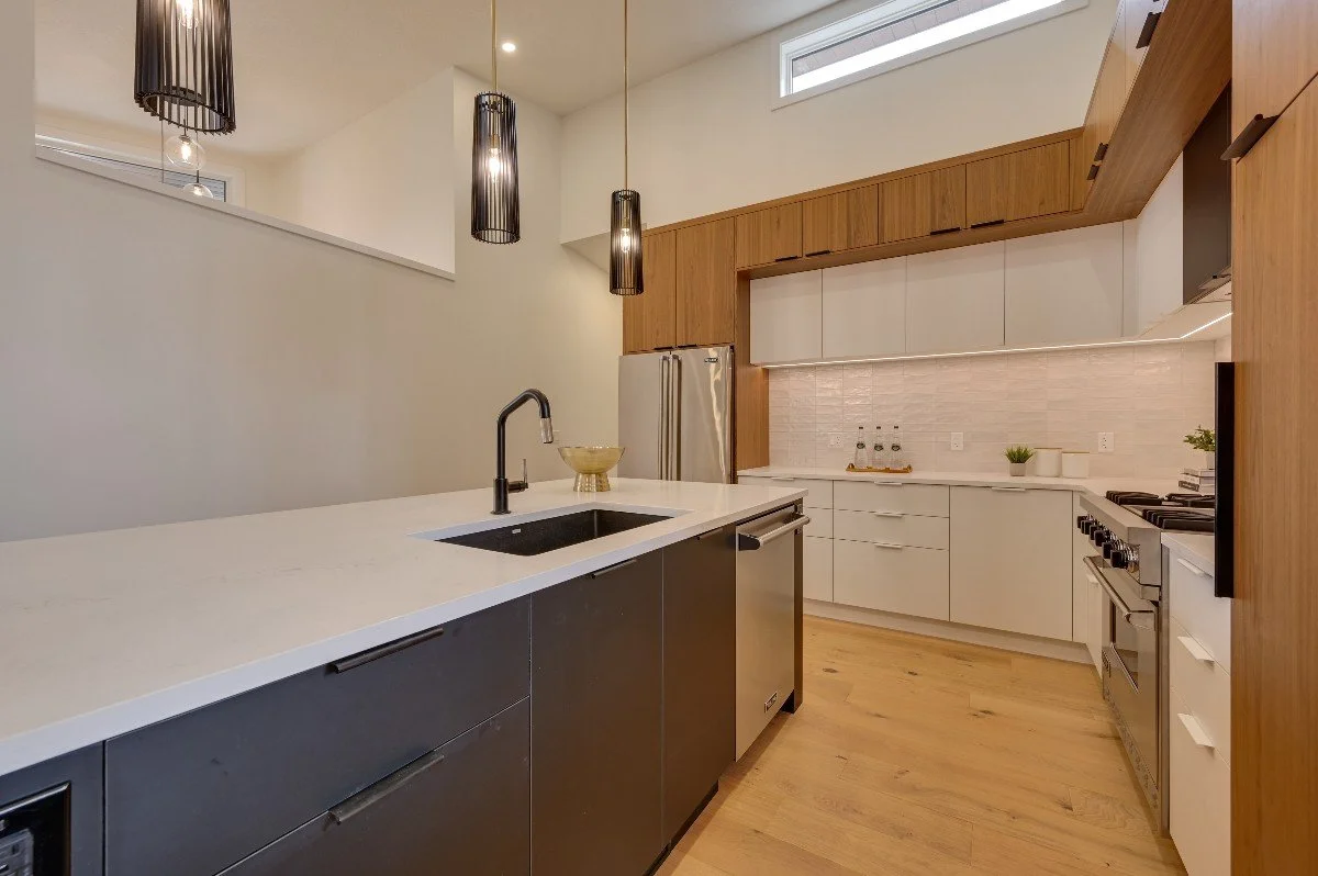 Modern kitchen with white and wood cabinets, black pendant lights, stainless steel appliances, a black sink with a black faucet, and a light wood floor.