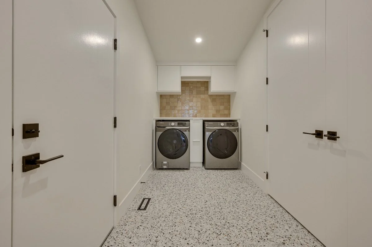Laundry room with a washer and dryer side by side, white cabinets above, beige tiled backsplash, and white walls.