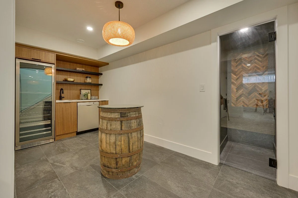 A small kitchenette with a mini fridge, wine cooler, wooden shelves, and a wooden barrel used as a table, with a pendant light hanging above.
