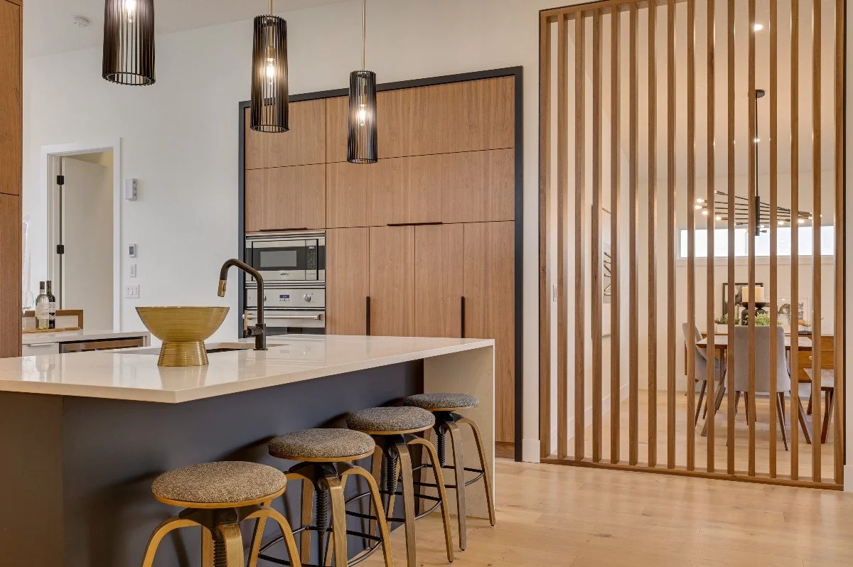 Modern kitchen with an island and three barstools, wooden cabinetry, pendant lighting, and a separate dining area visible through a wooden slat partition.