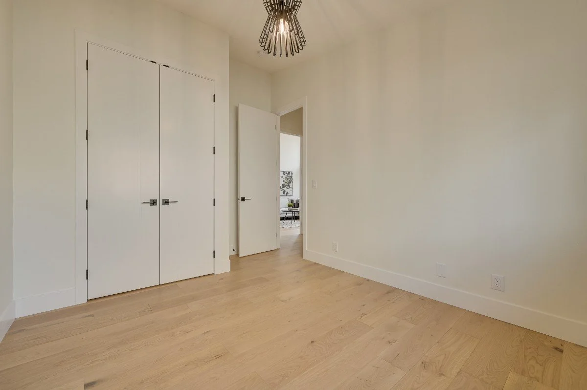 Empty room with white walls, light hardwood floors, a closed double closet door, an open door to another room, and a modern chandelier.