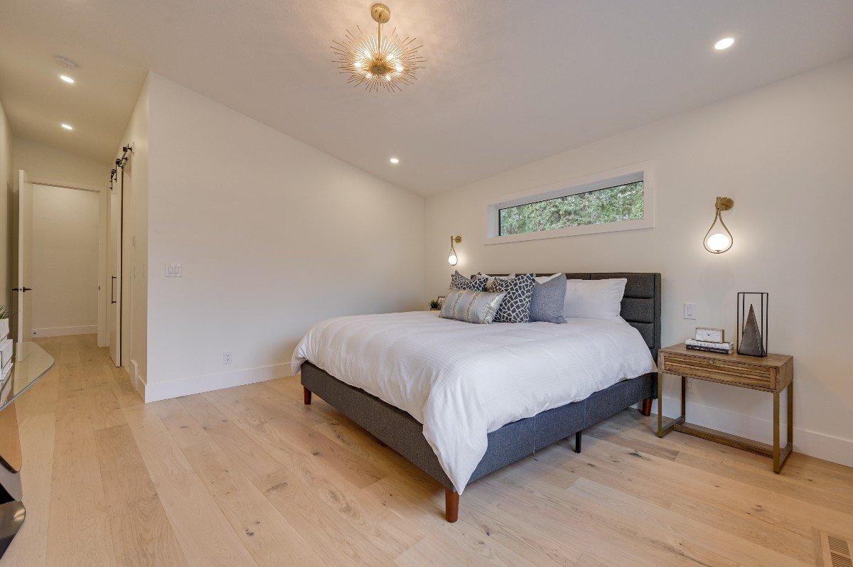 Modern bedroom with a gray bed frame, white bedding, decorative pillows, a wooden nightstand with a lamp and decor, a horizontal window near the ceiling, and a gold chandelier