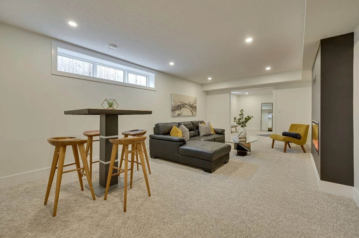 Living room with beige carpet, black sectional sofa with yellow and gray pillows, yellow armchair, coffee table with decorative items, wall-mounted fireplace, framed artwork on wall, slim horizontal window high on wall, bar table with four wooden sto