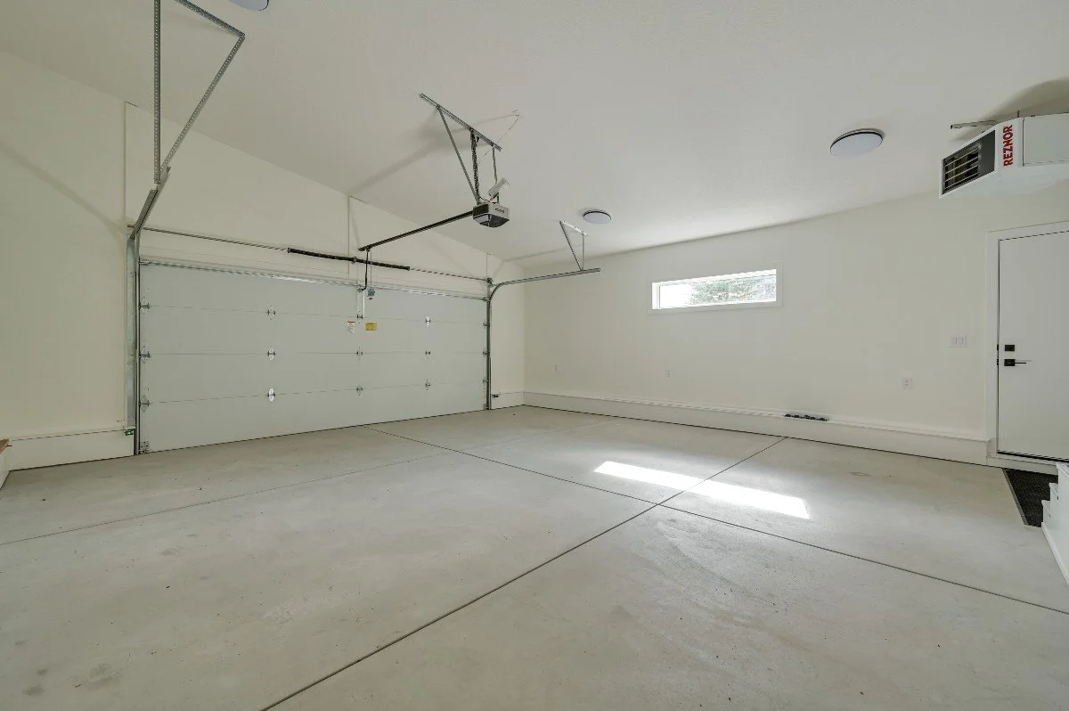 An empty residential garage with a closed white overhead door, a small horizontal window, a wall-mounted HVAC unit, and a concrete floor.