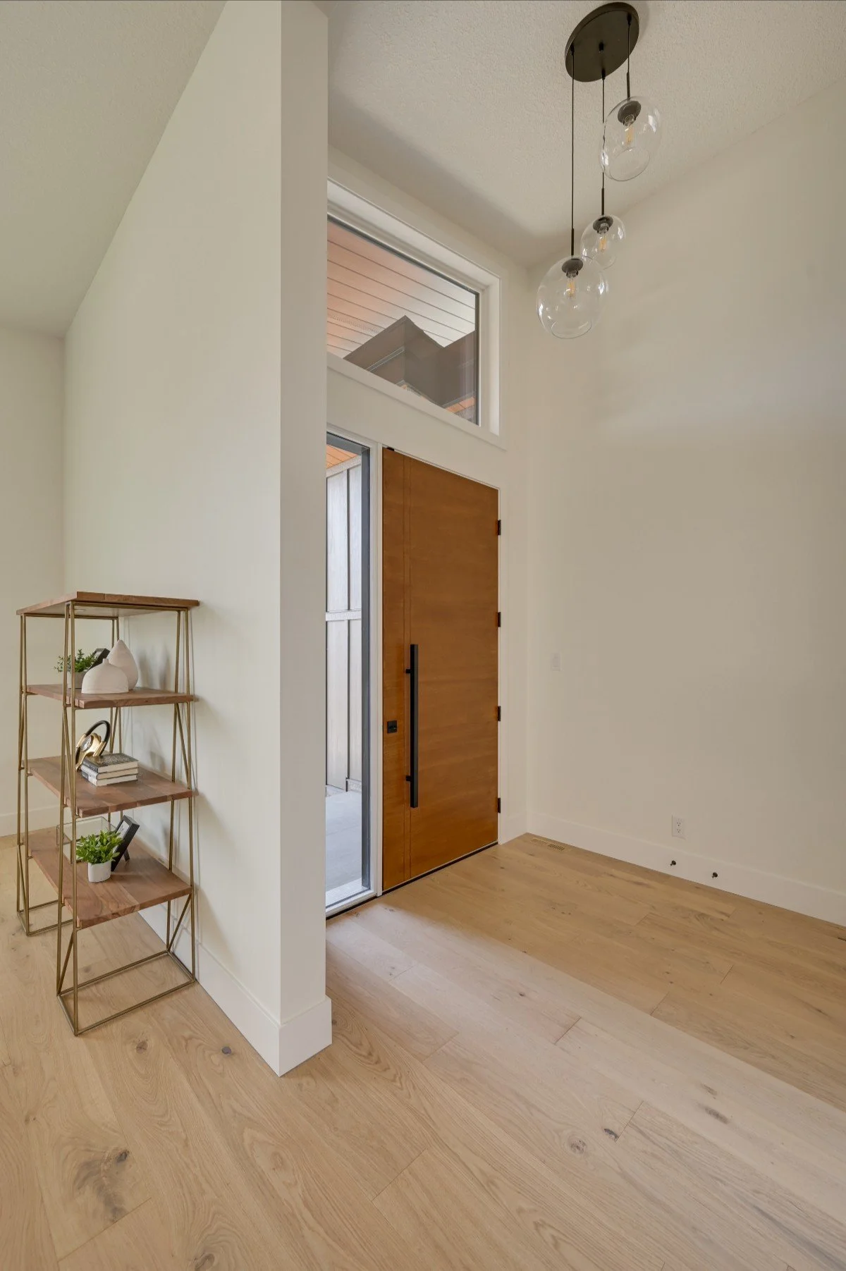 Modern entryway with wooden door, large window, and gold metal shelf with decor items, hardwood floor, and hanging light fixtures.