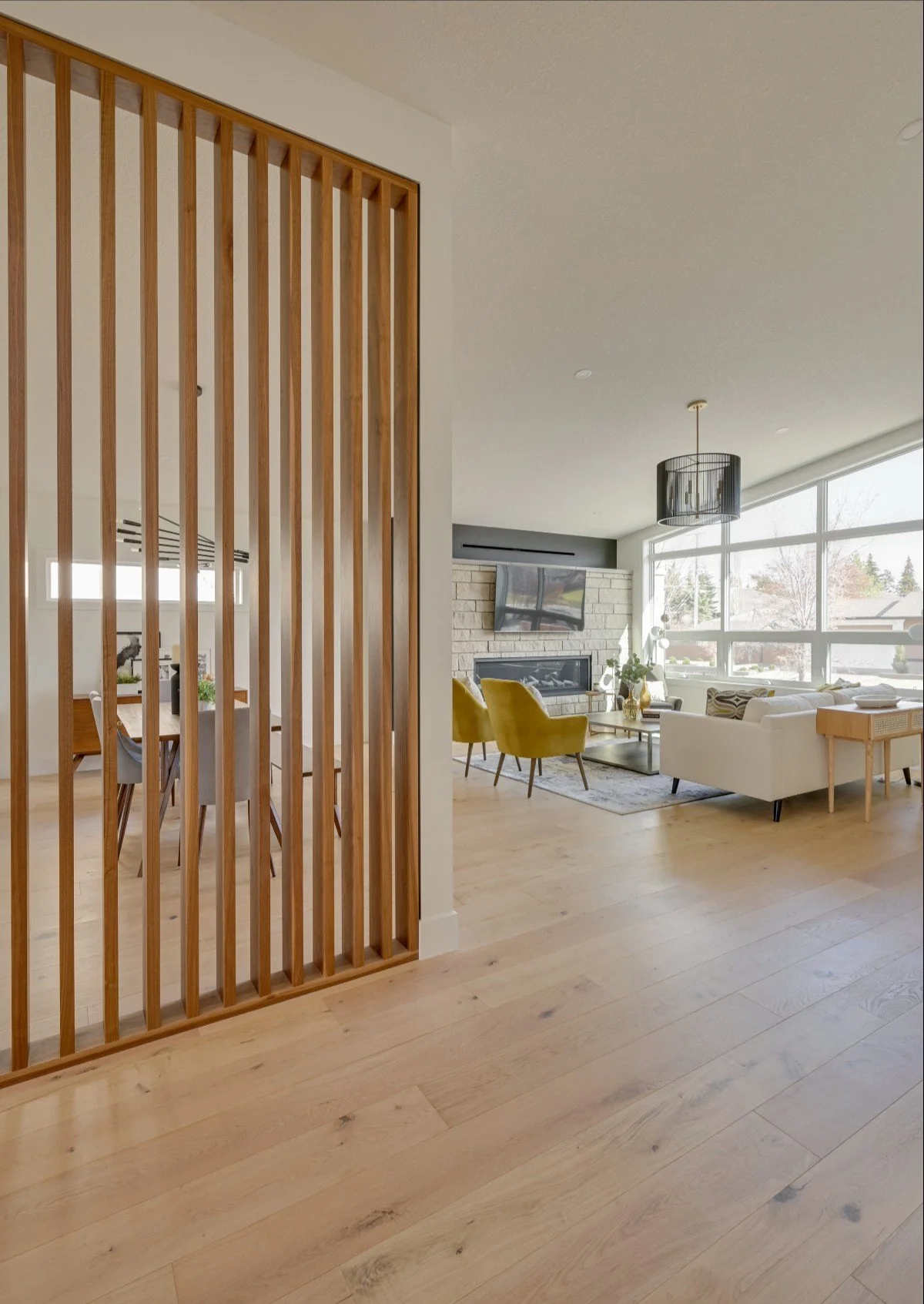 A modern living room with large windows, a white sofa, two yellow armchairs, a stone fireplace, and a ceiling light fixture. A wooden slat partition separates this space from the dining area.