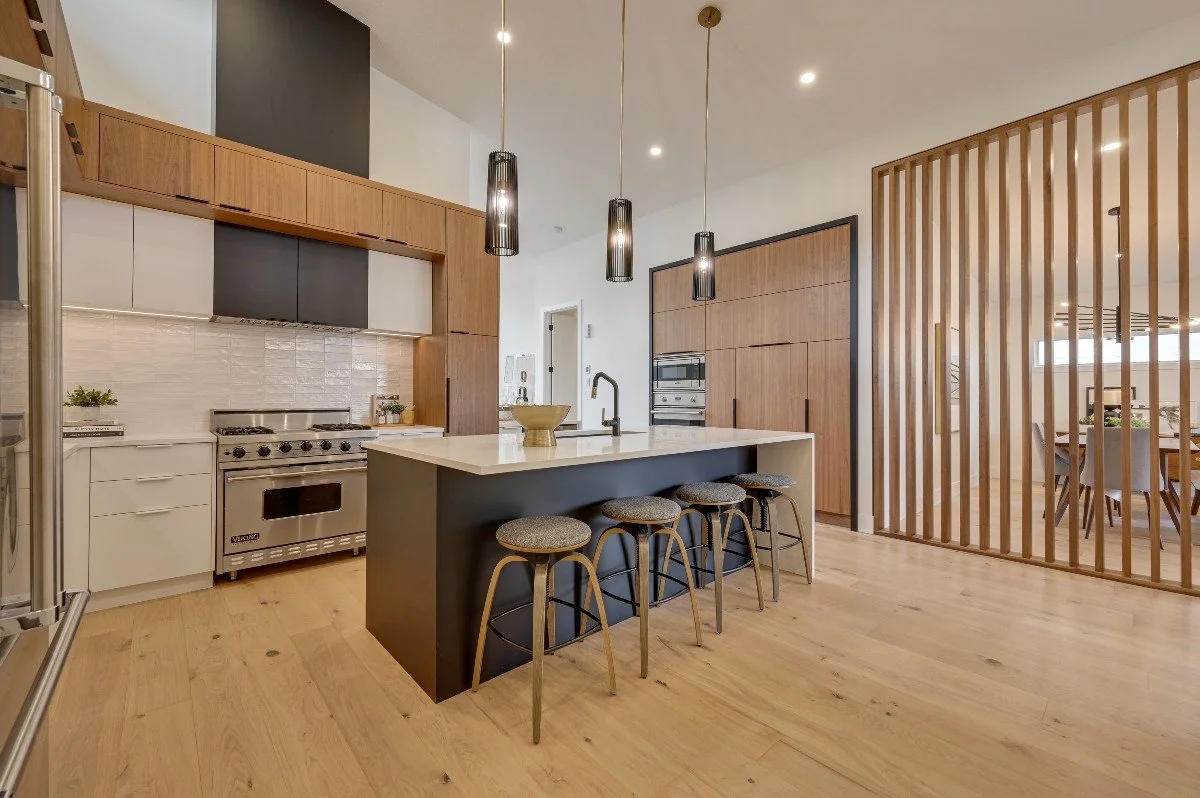 Modern kitchen with white and dark cabinets, stainless steel appliance, island with a sink and four stools, wood flooring, vertical wood slat divider, dining area in the background.