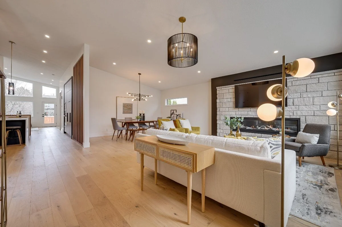 Open-concept living room and dining area in a modern home with light wood flooring, a stone fireplace, and stylish furniture including a white sofa, gray armchair, and a wooden side table, illuminated by contemporary lighting fixtures.