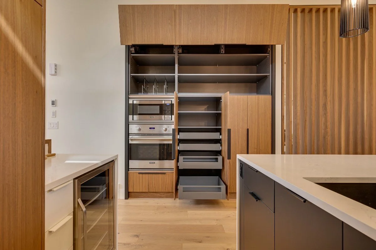 Modern kitchen with wooden cabinets, built-in stainless steel oven and microwave, open cabinet doors revealing pull-out drawers, white countertops, and a wood-paneled wall.