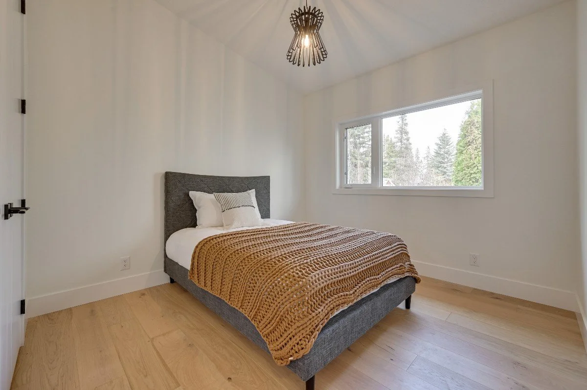 Empty bedroom with a bed, white linens, gray headboard, and a brown chunky knit blanket. There is a window showing trees outside, a black and brass chandelier, and a wooden floor.