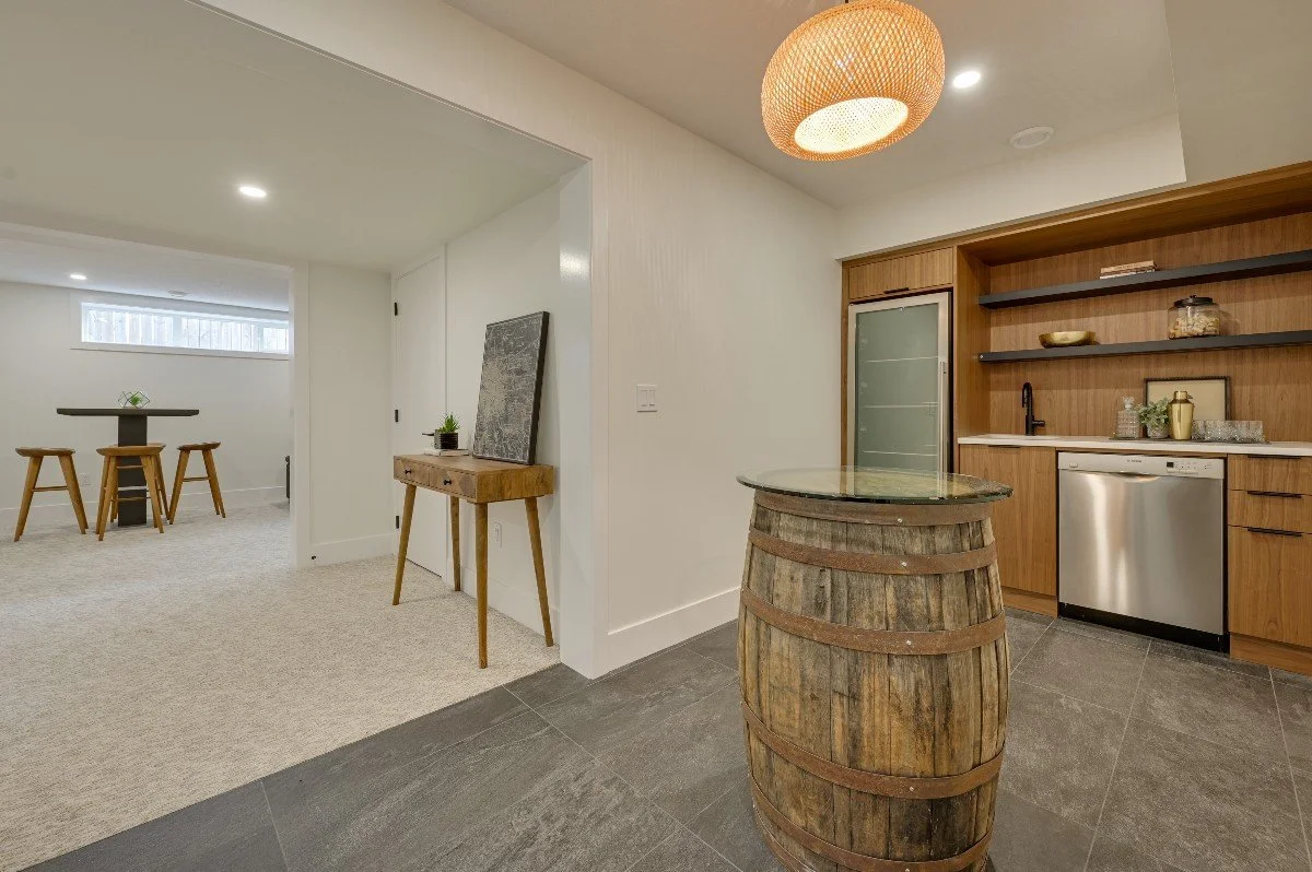 A room with a rustic wooden barrel used as a table, modern kitchen cabinets with open shelves, a dishwasher, and a wooden door, with a decorative ceiling light and minimal decor.