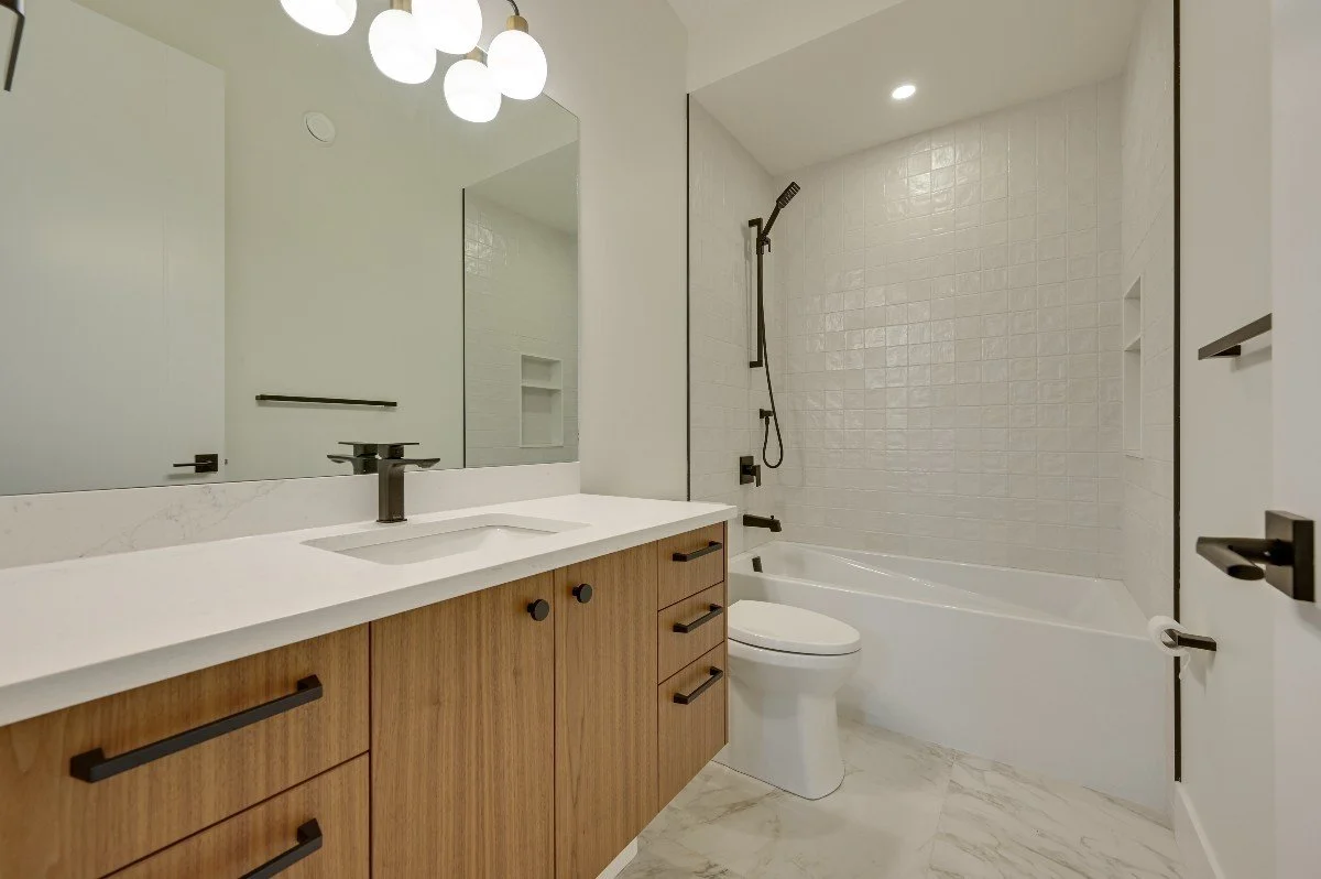 Modern bathroom with a wooden vanity, white sink, black faucet, large mirror, white tiled bathtub with a shower, and minimal black hardware.