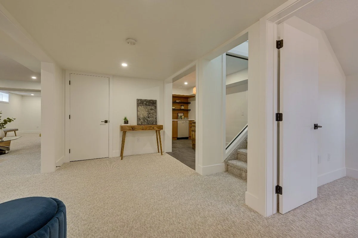 Empty basement room with beige carpet, white walls, and stairs to upper floor. Small table with potted plant and artwork, open doorway to kitchen, and partially visible door to closet or storage space.