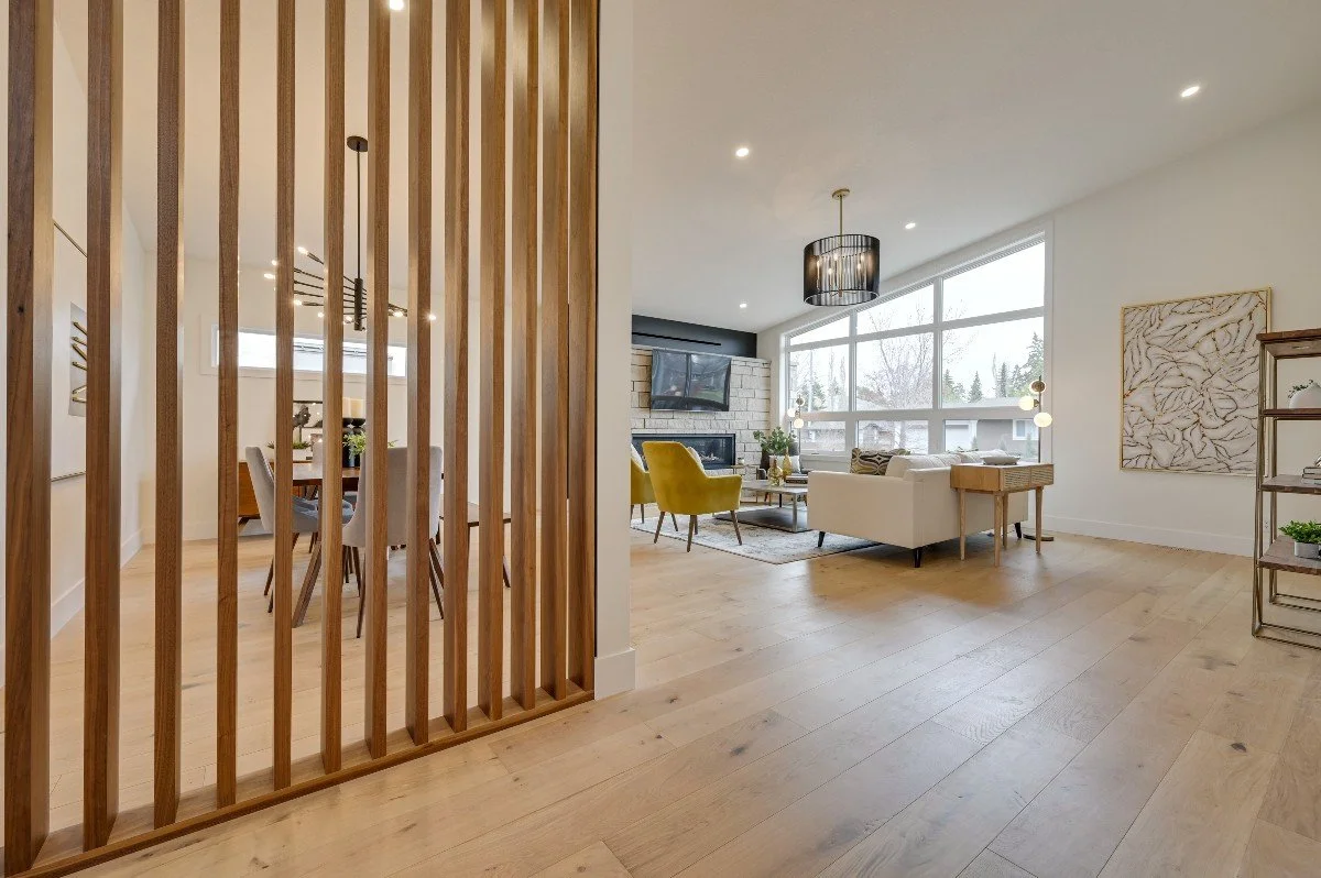 A spacious, modern living room with large windows, light-colored hardwood floors, a white sofa, yellow armchairs, a fireplace, and contemporary artwork.